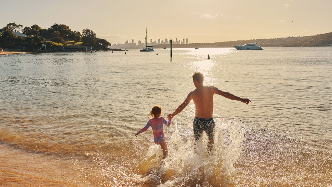 Parent and child splashing into the calm water at Camp Cove with the city skyline beyond