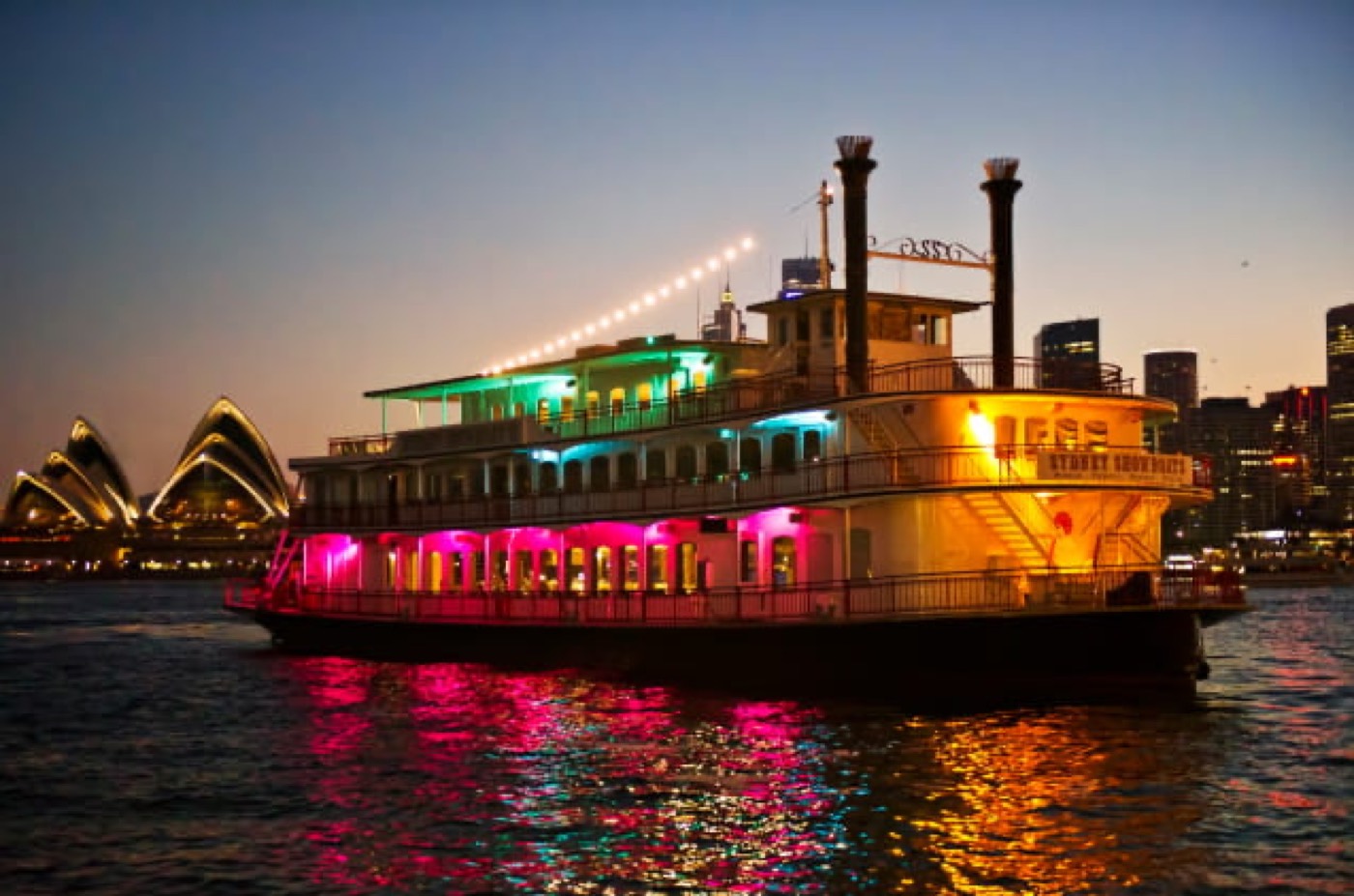 The Sydney Showboat paddlewheeler lit up at dusk with the Opera House behind