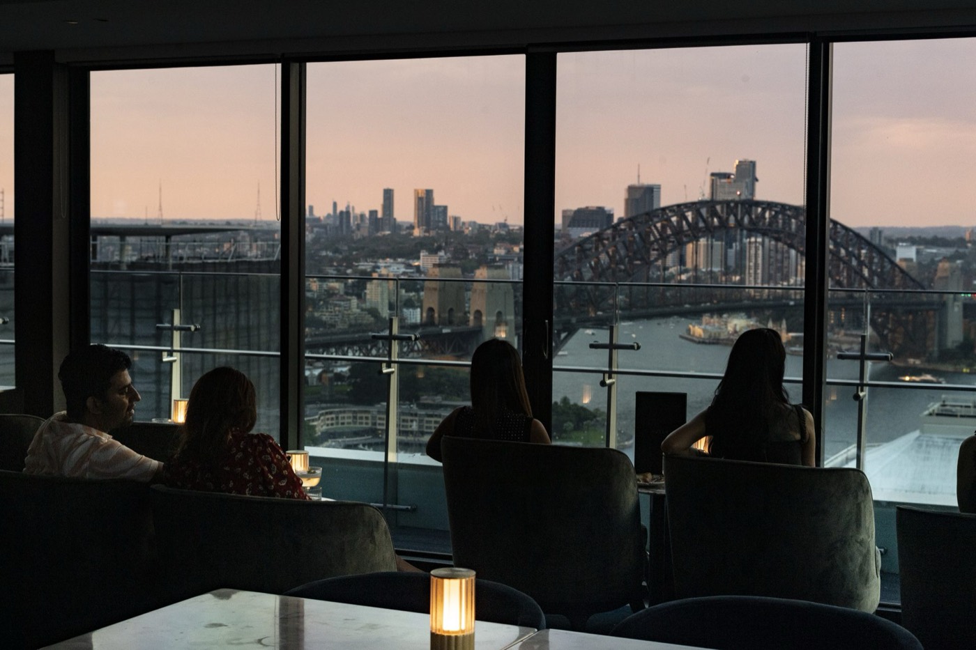 Guests at Aster looking through floor-to-ceiling windows at the Harbour Bridge at dusk