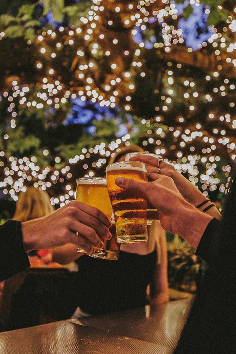 Friends clinking beers under fairy-lit branches at The Oaks