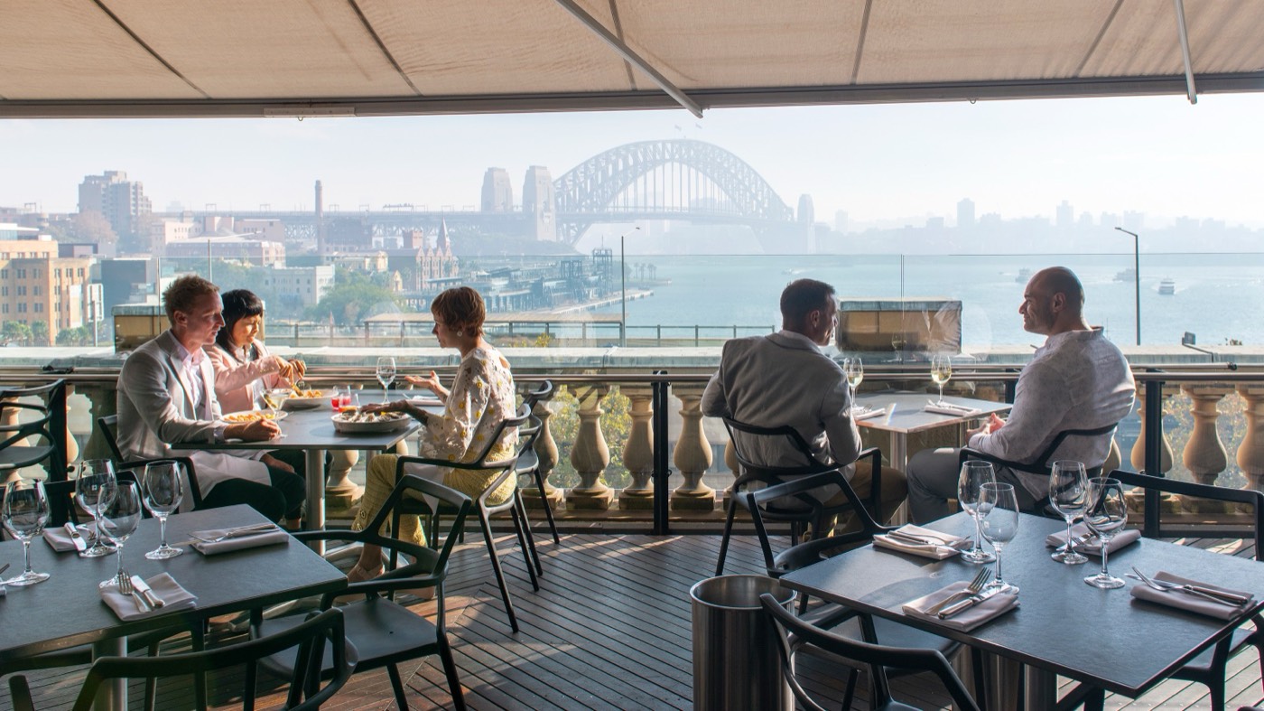 Diners at a harbour-view restaurant terrace with the Harbour Bridge in the distance