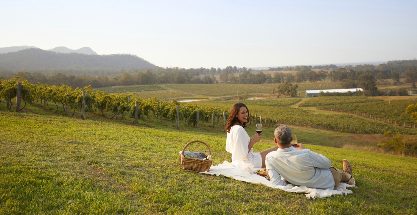 Couple picnicking with wine on a grassy slope above Hunter Valley vineyards