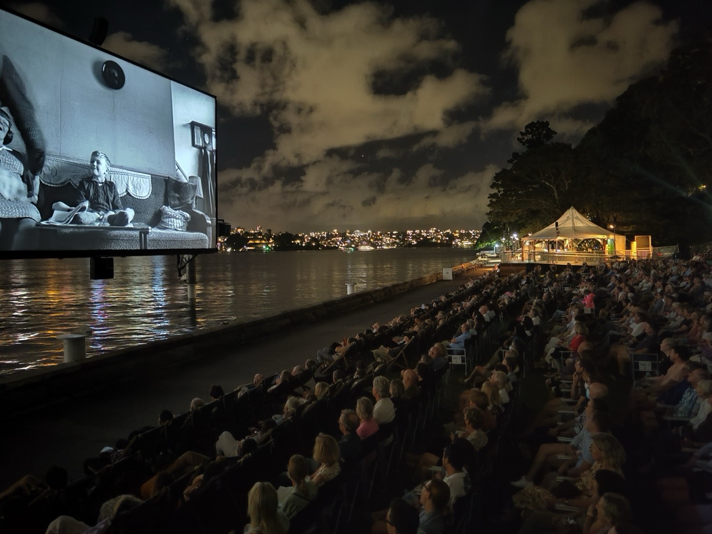 Audience watching a film on a big outdoor screen beside Sydney Harbour at night
