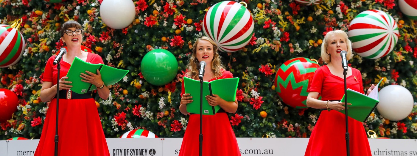 Three carollers in red dresses singing in front of the decorated Martin Place tree