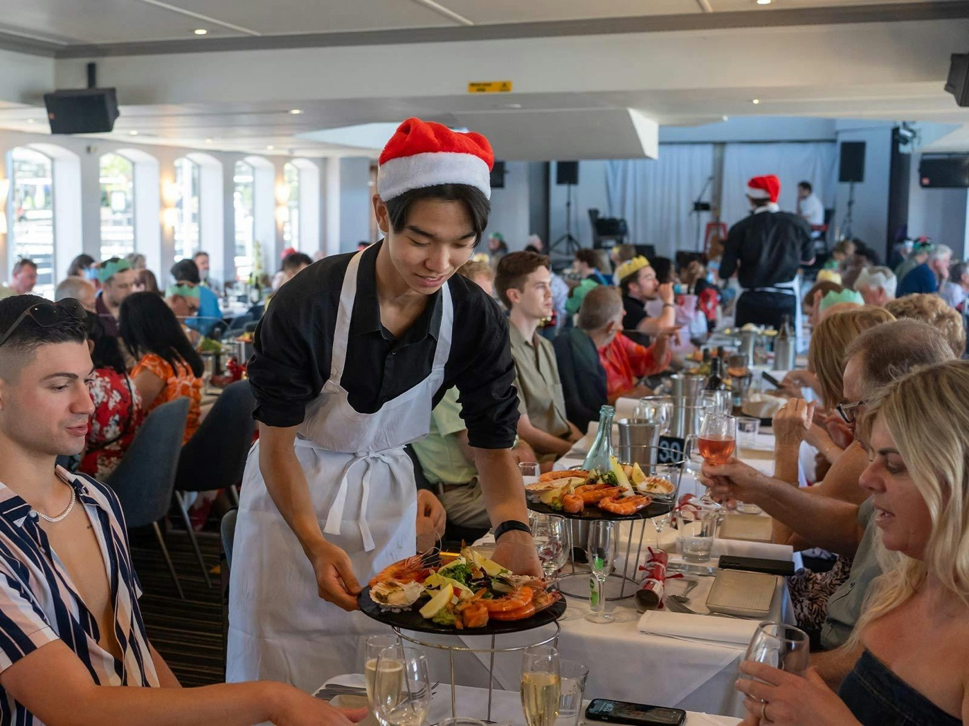 A waiter in a Santa hat serving seafood platters to guests at a Christmas lunch cruise