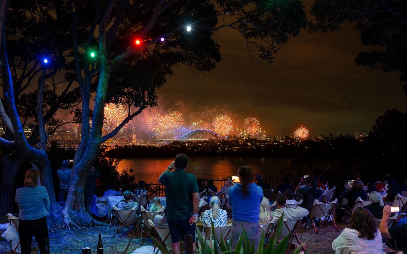 Guests under festoon lights at Taronga Zoo watching fireworks across the harbour