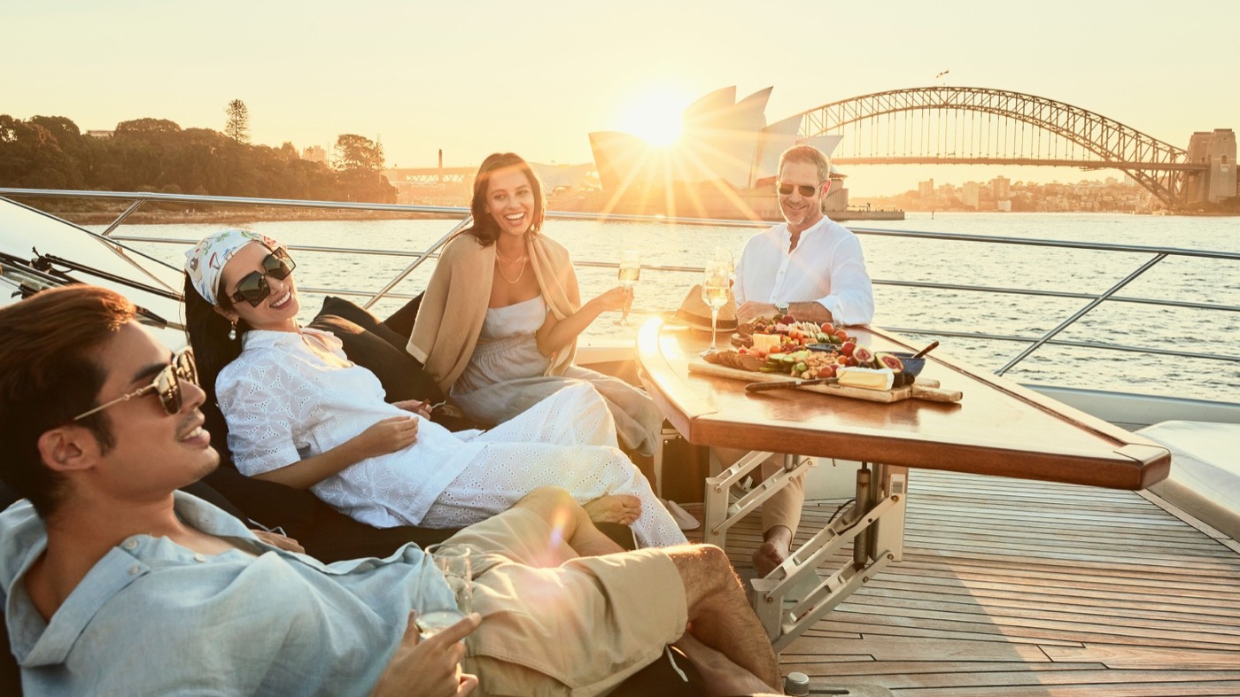 Friends relaxing on a yacht deck at sunset with the Opera House and Harbour Bridge behind