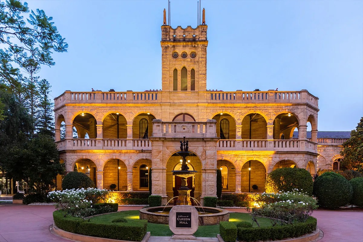 Curzon Hall's sandstone facade and tower lit up at dusk behind a fountain and rose garden